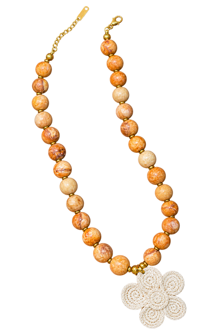 A beaded necklace featuring round, marbled orange and beige stone beads separated by small gold spacer beads, with a gold clasp and extender chain on one side and a white crocheted flower-shaped charm on the other, displayed on a solid white background.